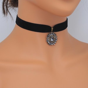 May include: A black velvet choker necklace with a decorative pendant. The pendant is an oval shape with a central, square-cut, gray gemstone surrounded by smaller gray stones. The metal setting has a gold tone.