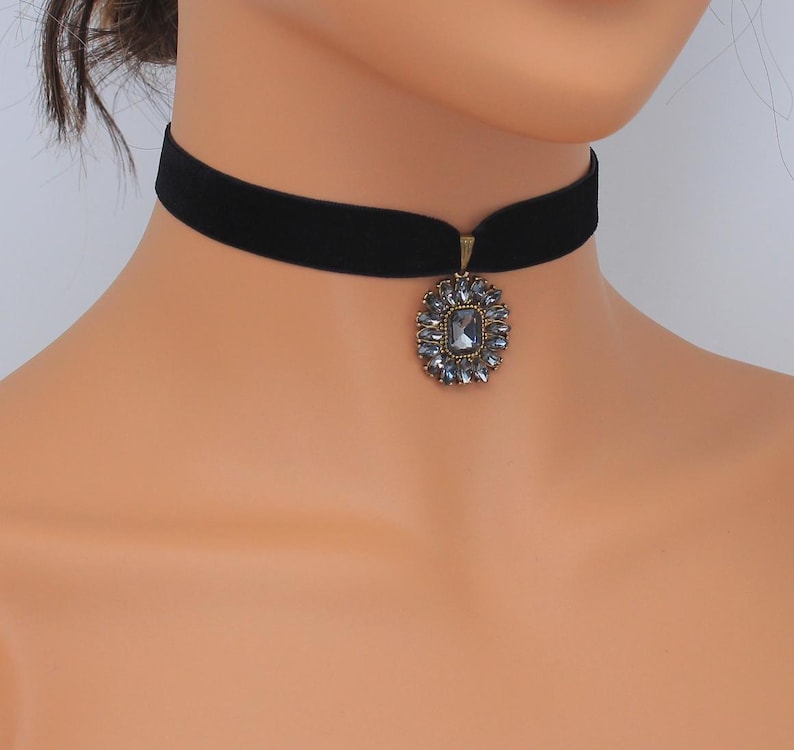May include: A black velvet choker necklace with a decorative pendant. The pendant is an oval shape with a central, square-cut, gray gemstone surrounded by smaller gray stones. The metal setting has a gold tone.