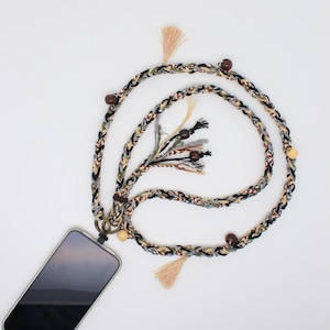 Op de afbeelding: Een gevlochten telefoonlanyard in een neutraal kleurenpalet van zwart, beige en grijs, geaccentueerd met houten kralen en kwastjes. De lanyard is bevestigd aan een smartphone, waardoor een stijlvol en functioneel accessoire ontstaat.
