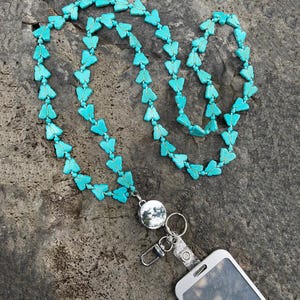 Puede incluir: Cordón con cuentas de mariposa turquesa con un carrete retráctil plateado y un clip para teléfono plateado. El cordón está hecho de pequeñas cuentas turquesas en forma de mariposa. Sobre una superficie gris texturizada.