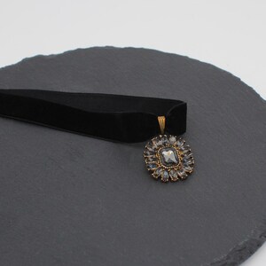 May include: A black velvet choker necklace with a square, gray gemstone pendant surrounded by smaller, clear stones. The pendant has a gold-colored metal frame. The necklace is displayed on a dark gray, textured surface.