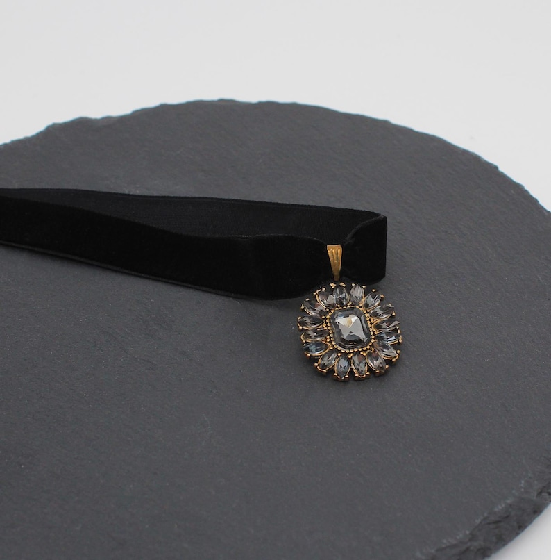 May include: A black velvet choker necklace with a square, gray gemstone pendant surrounded by smaller, clear stones. The pendant has a gold-colored metal frame. The necklace is displayed on a dark gray, textured surface.