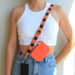 Puede incluir: Un bolso bandolera naranja con una correa a juego con lunares negros. El bolso incluye una pequeña bolsa naranja y una cadena dorada. La correa está sujeta a un cierre dorado. Un smartphone está sujeto al cierre.