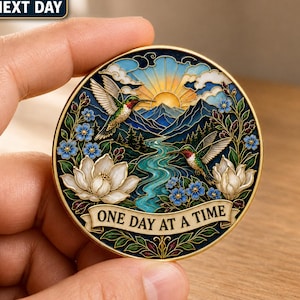 Può includere: Un medaglione circolare con bordo dorato che presenta una colorata illustrazione in smalto di due colibrì, fiori di magnolia bianchi, nontiscordardime blu, un fiume tortuoso, montagne e un sole. Un banner in basso riporta la scritta ONE DAY AT A TIME.