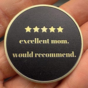 May include: A round, black coin with a gold border. The coin features five gold stars above the words "excellent mom. would recommend." The text and stars are in a gold color, contrasting against the black background.