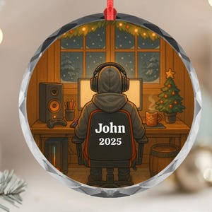 May include: A round, clear Christmas ornament with a red ribbon. The ornament features a cartoon illustration of a person wearing headphones, sitting at a desk with a computer. The text "John 2025" is on the chair.
