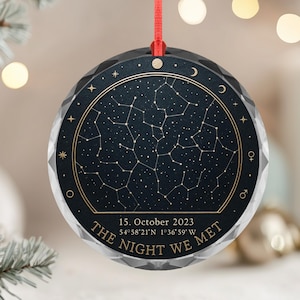 Puede incluir: Un adorno redondo y facetado con un fondo azul oscuro y un diseño de constelación dorada. El texto "THE NIGHT WE MET" y la fecha "15. October 2023" están impresos en el adorno. Se adjunta una cinta roja para colgarlo.