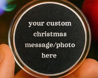 May include: A clear, round Christmas ornament with a black center. The text "your custom christmas message/photo here" is printed in white. The ornament is held in a hand, with a blurred Christmas tree and ornaments in the background.