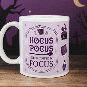 Op de afbeelding: Witte keramische mok met een paars ontwerp. De mok heeft de tekst "Hocus Pocus I Need Coffee To Focus" en illustraties van een drankfles, ketel, spreukenboek en bezem. De achtergrond toont een Halloween-scène met een maan.