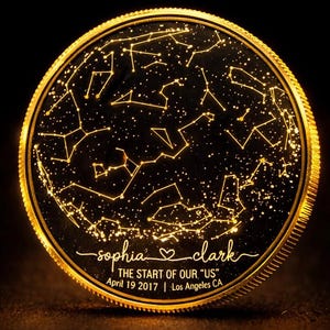 Puede incluir: Un recuerdo redondo de color dorado que presenta un mapa de constelaciones con estrellas iluminadas. El texto dice "sophia ♡ clark", "THE START OF OUR 'US'" y "April 19 2017 | Los Angeles CA". El diseño tiene un fondo oscuro con un borde dorado.