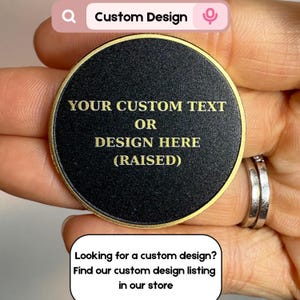 May include: A black, round custom design item with gold lettering and a gold border. The text reads "YOUR CUSTOM TEXT OR DESIGN HERE (RAISED)". The item is held in a hand. The top of the image has the text "Custom Design".
