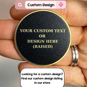 Pode incluir: Um item redondo preto com um design personalizado e letras douradas. O texto diz "YOUR CUSTOM TEXT OR DESIGN HERE (RAISED)". O item &eacute; segurado em uma m&atilde;o. A imagem tamb&eacute;m inclui o texto "Looking for a custom design? Find our custom design listing in our store."