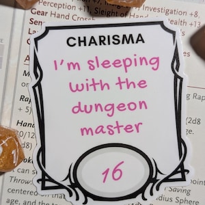 May include: A white sticker with a black border, featuring the word "CHARISMA" at the top. The text "I'm sleeping with the dungeon master" is in pink. A pink "16" is in an oval at the bottom. Gold dice are in the background.