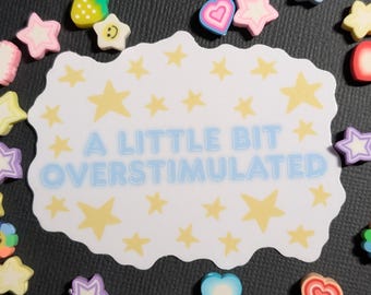 May include: A white rectangular sticker with wavy edges featuring the text A LITTLE BIT OVERSTIMULATED in light blue letters surrounded by yellow stars. The sticker is scattered with small colorful erasers shaped like stars, hearts, and flowers.