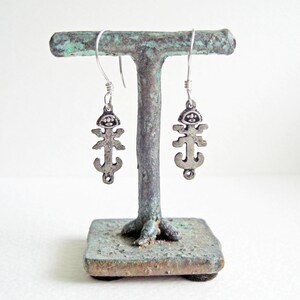 Puede incluir: Un par de pendientes colgantes plateados. Cada pendiente presenta una figura estilizada con cabeza, brazos y una sección inferior que se asemeja a un ancla. Los pendientes están suspendidos de ganchos plateados. Los pendientes se exhiben en un soporte de metal envejecido.