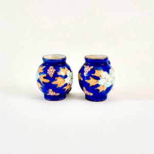以下が含まれることがあります： 丸みを帯びたコバルトブルーの小さな花瓶2つ。 金色の縁と取っ手があり、オレンジと白の花と葉が描かれています。 背景は無地の白です。
