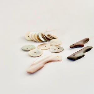 May include: A collection of various buttons in neutral tones. The assortment includes round, oval, and elongated shapes, with a mix of colors from cream to brown. Some buttons have two holes, while others have a single hole.