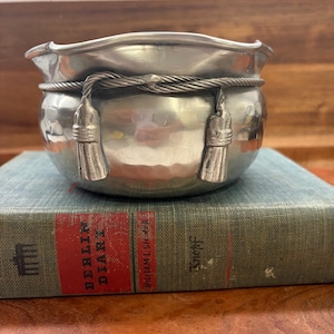 Op de afbeelding: Een kleine, zilverkleurige metalen kom met een getextureerd oppervlak en een gedraaid touwontwerp in het midden. Twee kwasten hangen aan het touw. De kom staat op een vintage boek met de titel "Berlin Diary".