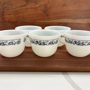 May include: Five white ceramic teacups with a blue floral design around the rim. The cups are arranged on a wooden cutting board. The teacups have a handle and a rounded shape. The background includes a wooden tray and a white surface.