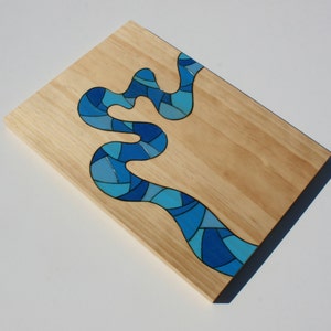 May include: A rectangular wooden serving board with a river design. The river is composed of various shades of blue, creating a mosaic effect. The board has a natural wood grain finish and a dark blue border.