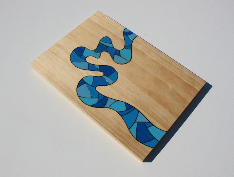 May include: A rectangular wooden serving board with a river design. The river is composed of various shades of blue, creating a mosaic effect. The board has a natural wood grain finish and a dark blue border.