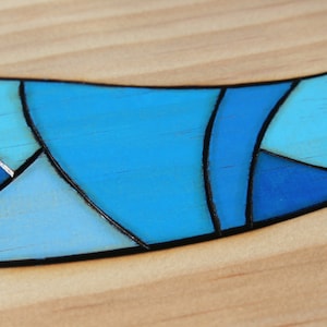 May include: Close-up of a wooden surface with a curved, mosaic-like design. The design features various shades of blue, outlined in black, creating a stained-glass effect. The background is light-colored wood.