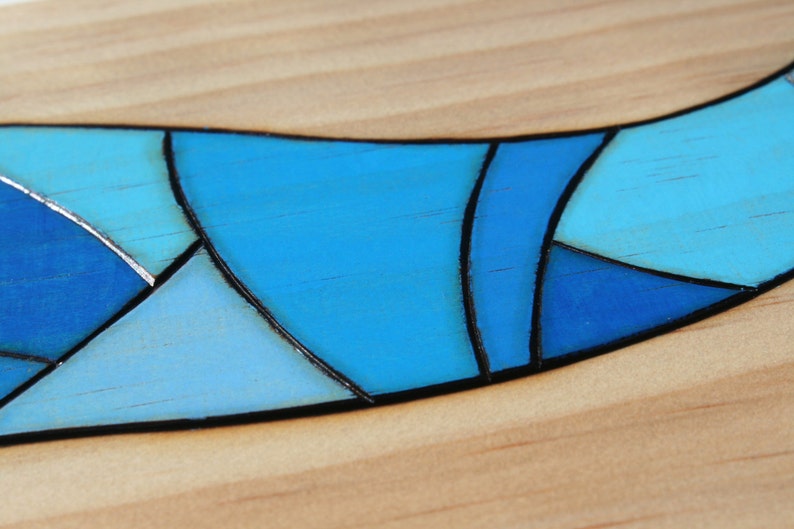May include: Close-up of a wooden surface with a curved, mosaic-like design. The design features various shades of blue, outlined in black, creating a stained-glass effect. The background is light-colored wood.