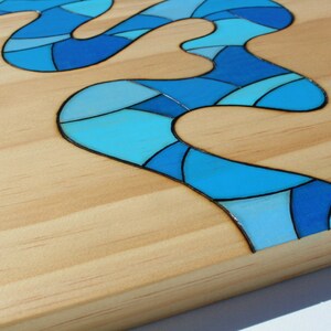 May include: Wooden art piece featuring a flowing river design in shades of blue. The river is composed of geometric shapes and is set against a light-colored wood background. The artwork has a glossy finish and a modern aesthetic.