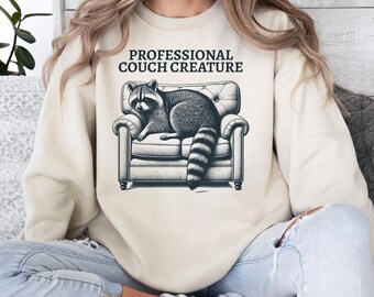 May include: Cream-colored sweatshirt featuring a graphic of a raccoon lounging on a small couch. The text "PROFESSIONAL COUCH CREATURE" is printed above the graphic in black. The sweatshirt is paired with blue jeans.