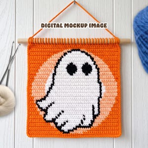 Könnte beinhalten: Ein quadratischer Häkel-Wandbehang mit einem weißen Geist mit schwarzen Augen, zentriert auf einem hellorangen Kreis vor einem leuchtend orangen Hintergrund. Das Stück hängt an einem Holzstab mit einer orangen Garnaufhängung. Text: DIGITAL MOCKUP IMAGE.
