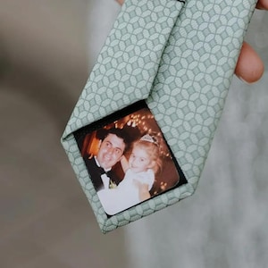 May include: A light green tie with a geometric pattern. The inside of the tie features a photograph of a man and a young girl. The tie is held by a person.