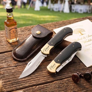 May include: Two folding pocket knives with black handles and brass bolsters, one open and one closed, featuring the names Robert and Oliver engraved on the brass. Includes a brown leather sheath, a small bottle of liquor, and a wedding invitation.