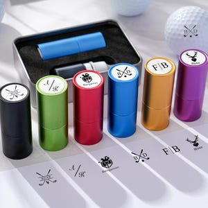 May include: A collection of personalized golf ball stamps in various colors, including black, green, red, blue, gold, and purple. Each stamp has a white top with a unique design and initials. A golf ball and a metal case with a blue item are also visible.