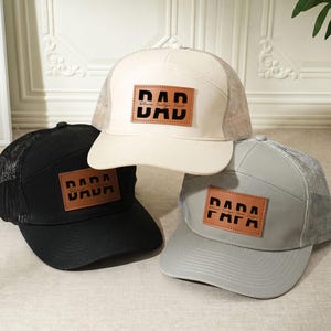 May include: Three trucker hats in black, cream, and gray. Each hat has a mesh back and a square, brown leather patch with the word "DAD" or "PAPA" in bold, black letters. The cream hat has the text "Believe Create Find" below the word "DAD".
