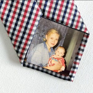 Könnte beinhalten: Eine Krawatte mit Fotoeinsatz. Die Krawatte hat ein rot-weiß-blaues Karomuster auf der Vorderseite und ein Foto einer Frau und eines Babys. Die Krawatte ist aus gewebtem Stoff und wird auf einer weißen Oberfläche präsentiert.
