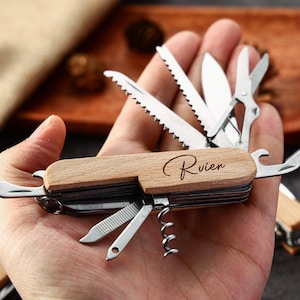 May include: A multi-tool with a wooden handle and various stainless steel tools, including a saw, scissors, and bottle opener. The wooden handle is engraved with the name "Rivien". The tool is held in a hand.