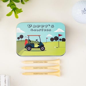 Op de afbeelding: Een golfcadeauset met een blik met een golfbaan en de tekst "DADDY'S GOLFTEES". Inclusief drie houten tees met dezelfde tekst en twee golfballen. Het blik is ongeveer 10 cm lang.