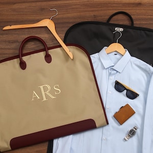 Puede incluir: Una camisa azul claro, gafas de sol, una cartera marrón y un reloj plateado se muestran con una funda de ropa negra y una funda de ropa beige con las iniciales "ARS" bordadas. Se incluye una percha de madera.