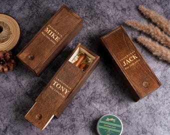 May include: Three wooden boxes with sliding lids, engraved with names and dates, likely for groomsmen gifts. One box is open, revealing cigars and filler. The text on the boxes reads "Groomsman" and names like "Mike", "Jack", and "Tony".