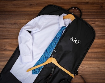 Puede incluir: Una camisa blanca y una corbata azul con estampado de cachemira se muestran en una funda de ropa negra. La bolsa tiene las iniciales "ARS" impresas. Una percha de madera sostiene la camisa y la corbata, lo que sugiere ropa formal.