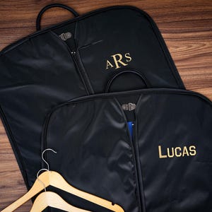 May include: Two black garment bags with gold embroidered initials and names. One bag has "ARS" embroidered, the other has "LUCAS". Wooden hangers are visible in the foreground. The bags are on a wooden surface.