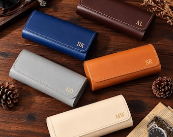 Puede incluir: Cinco estuches rectangulares para gafas en colores marrón oscuro, azul, gris, tostado y crema. Cada estuche cuenta con un cierre de solapa con costuras visibles e iniciales grabadas en oro: AL, BK, KD, SR y MW. Los estuches están sobre madera.