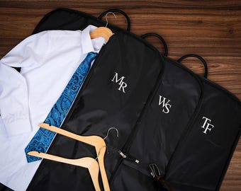 Puede incluir: Una camisa blanca y una corbata azul con estampado de cachemira descansan sobre una percha de madera. Se ven tres fundas de ropa negras con iniciales blancas. Las bolsas están sobre una superficie de madera.
