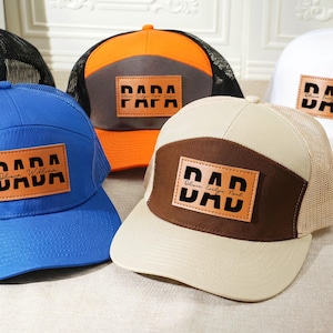 May include: A collection of trucker hats in various colors, including blue, orange, and white. Each hat features a leather patch with the word "DAD" or "PAPA" in bold, block letters. The hats have mesh backs and curved brims.
