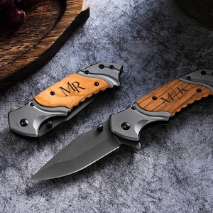May include: Two personalized folding knives with wooden handles and gray blades are displayed on a textured surface. The handles are engraved with initials, "MR" and "MK". The knives are closed, showing the wooden handle and the gray metal frame.