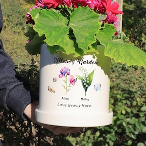 May include: White ceramic planter with a floral design and the text "Mom's Garden". The planter features two butterflies, a sweet pea flower, and a lily of the valley flower. The text "Addison" and "Mallory" are written below the flowers. The text "Love Grows Here" is written at the bottom of the planter.