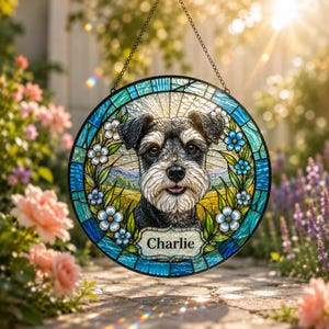 May include: A circular stained glass suncatcher featuring a portrait of a Schnauzer dog surrounded by blue flowers and green leaves. The piece includes a white rectangular plaque at the bottom with the name Charlie printed in black text.