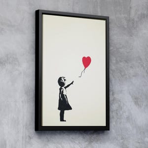 Op de afbeelding: Een zwart-wit sjabloondruk van een meisje dat naar een rode hartvormige ballon reikt. De print is ingelijst in een zwart frame.