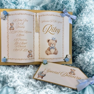 May include: A baby announcement card with a gold border, featuring a teddy bear illustration and the name "Riley." The card includes the text "A Baby Boy Is A Blessing" and "Welcome To The World." A smaller tag reads "Special Delivery."