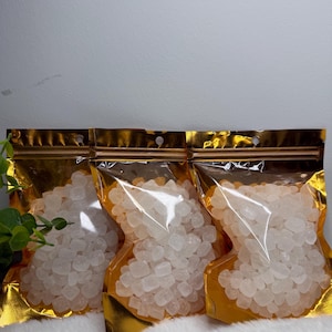 May include: Three clear plastic pouches filled with white, crystalline granules. The pouches have a gold-colored, metallic border and a zip closure. The background is a soft, white surface.